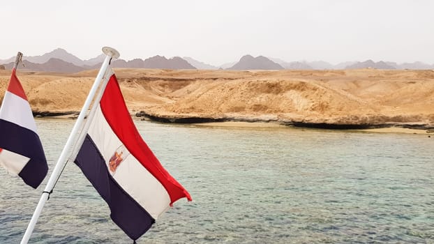The national flag flutters in the wind against the azure water of the Red Sea and the rocky sandy shore. Close-up. Landscape of the Red Sea. Excursion on a ship to Sharm El Sheikh