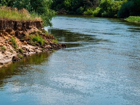 The course of the river along the steep bank. Water stream. Freshwater river Teterev. Steep coastline. Green leaves of trees and plants. Summer season. Natural landscape. Fishing place.