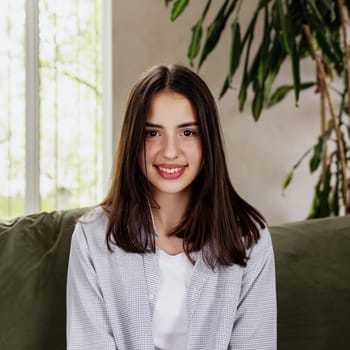 Beautiful smiling caucasian girl sitting at green sofa and watching at camera. Cute woman front view. Female smile, looking in photo camera. Online interview or meeting. Big lips. High quality photo