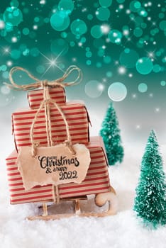 Sled With Christmas Gift And Label With English Calligraphy Merry Christmas And A Happy 2022. Snow With Christmas Tree And Green Sparkling Background