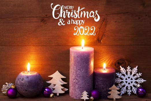 English Calligraphy Merry Christmas And Happy 2022. Purple Romantic Candle Light With Christmas Decoration. Brown Wooden Background