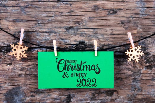 Green Label With English Calligraphy Merry Christmas And Happy 2022. Rope WIth Christmas Ornamen. Rustic Wooden Background