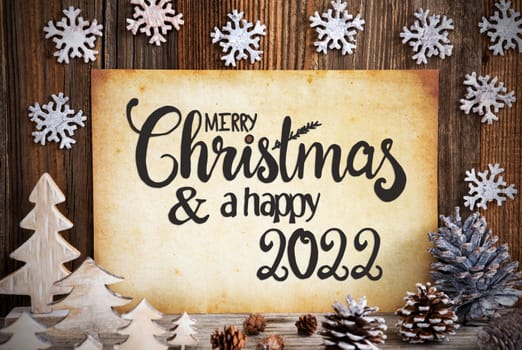 Old Paper With English Text Merry Christmas And Happy 2022. Christmas Decoration Like Tree, Fir Cone And Snowflakes. Brown Wooden Background
