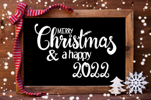 Chalkboard With English Calligraphy Merry Christmas And A Happy 2022. Christmas Decoration Like Tree And Bow. Wooden Background And Snowflakes