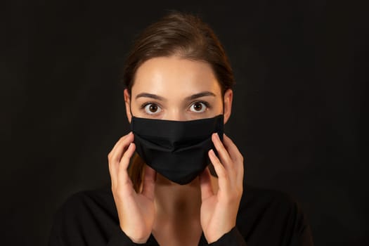 Attractive young woman in a black protective mask on dark background. Girl in a flu face protection touching her face against black backdrop. Covid-19 pandemic safety concern.