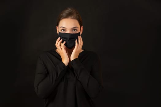 Attractive girl in black antiviral mask concerned about coronavirus prevention looking at the camera with copy space for text. Brunette young woman in studio touching her face.