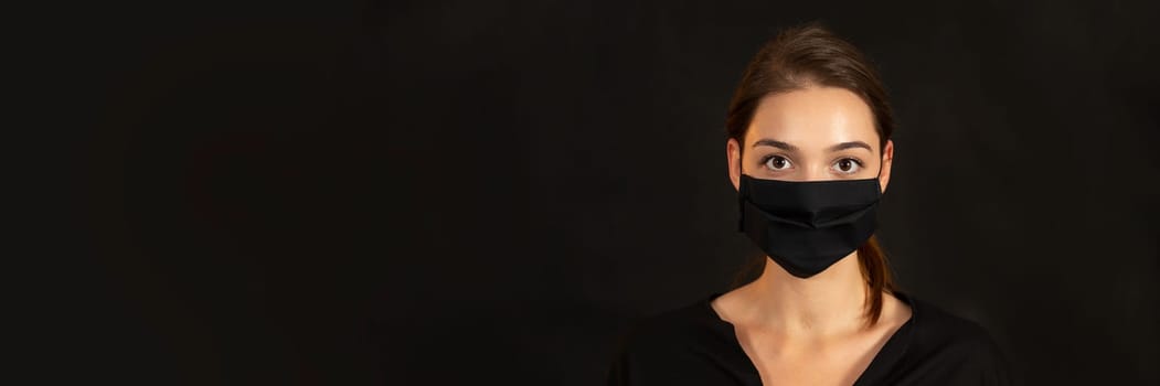 Horizontal head and shoulders portrait of a woman in protective mask amidst a Covid-19 pandemic. Studio headshot of attractive female against black backdrop with generous copyspace.