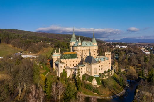 Popular tourist destination of Bojnice castle on a good weather day, with moat, greenery and forest around it. Medieval chateau from bird's eye view.