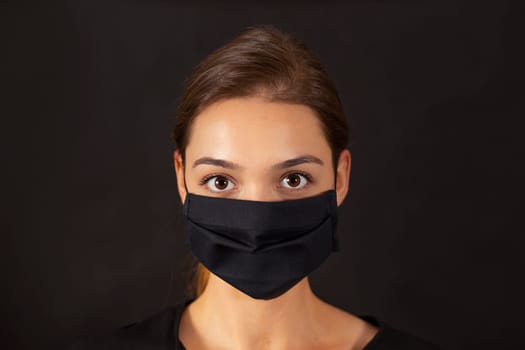 Studio close-up of a youthful girl wearing a black cloth face mask during a Covid-19 pandemic. Headshot portrait of a young woman in a protective medical equipment on dark background.