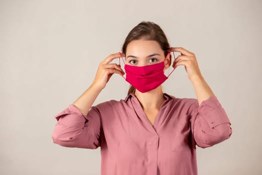 Youthful woman concerned about coronavirus putting on a red cloth mask to protect herself. Female nurse in the process of attaching a medical mask.