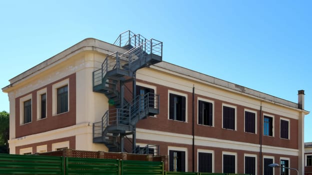 A well-preserved historic building in Civitavecchia, Italy, showcasing an external metal staircase. The building has a traditional design with numerous windows and a green fence.