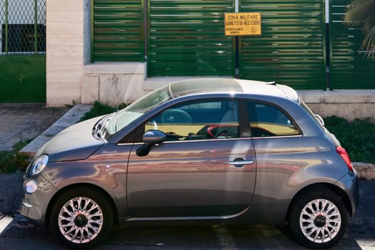Civitavecchia, Italy - January 26, 2025: A small car parked next to a green fence with a yellow military zone sign, highlighting the contrast between the civilian vehicle and the restricted area.
