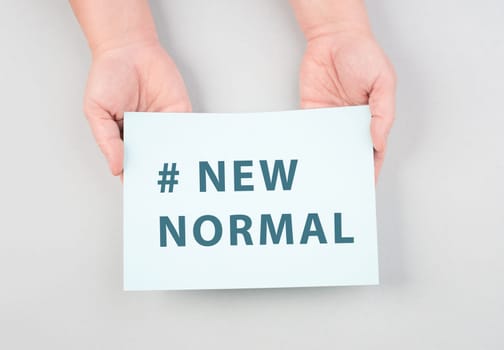 The words new normal are standing on a paper, hands holding the message, grey colored background, hashtag