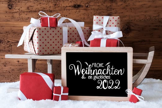 Blackboard With German Calligraphy Frohe Weihnachten Und Ein Glueckliches 2022 Means Merry Christmas And A Happy 2022. Sled With Christmas Gift And Presents. Wooden Background With Snow.
