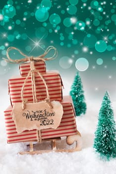 Sled With Christmas Gift And Label With German Frohe Weihnachten Und Ein Glueckliches 2022 Means Merry Christmas And A Happy 2022. Snow With Christmas Tree And Green Sparkling Background