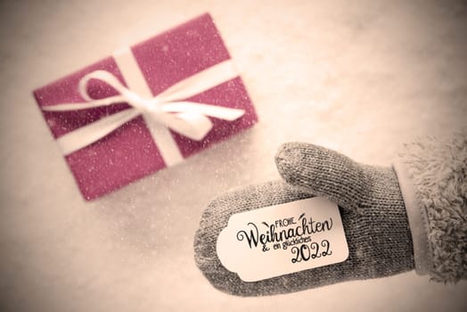 Label With German Calligraphy Frohe Weihnachten Und Ein Glueckliches 2022 Means Merry Christmas And A Happy 2022. Gray Glove With Pink Gift And Snow Background And Snowflakes