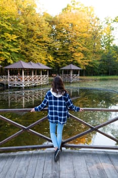 A young brunette woman in a plaid shirt stands with her back near the pond and enjoys the autumn warm day