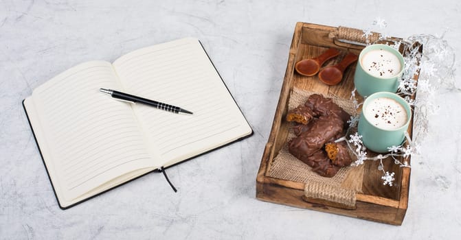 Coffee cups and chocolate coated gingerbread in a wooden basket, christmas season empty copy space, book with a pen