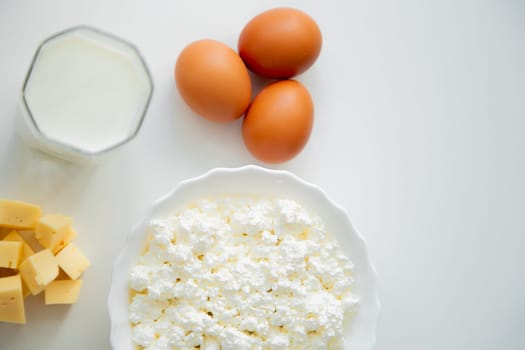 A white bowl with cottage cheese, eggs, diced cheese and a glass of milk stand on a white background. . High quality photo