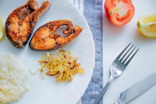 Fried fish with onions, rice and lemon lies on a plate, on a waffle towel and cutlery on a white background. High quality photo