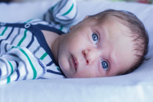 A small baby with blue eyes in a striped suit lies on a white pillow and looks slightly to the side of the frame. High quality photo