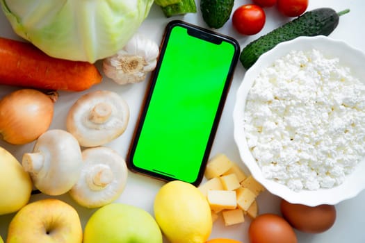 A phone with a green screen lies among fruits, vegetables and dairy products. High quality photo