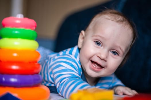 A little kid in a striped suit is lying on his tummy, next to him a children's pyramid and toys. High quality photo