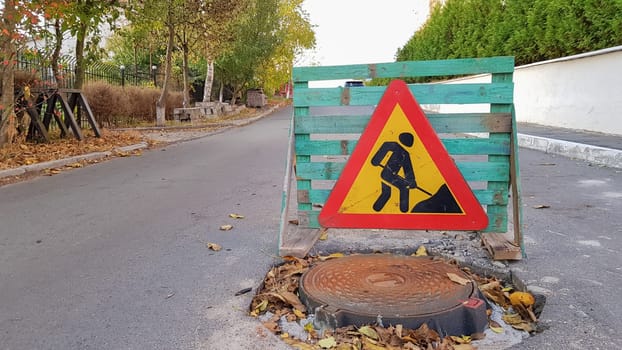 Road repair. Sewer manhole on the roadway. Triangular warning car drivers sign with a man with a yellow spade with red. Men at work. Traffic signs, travel restrictions