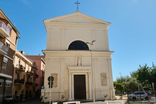 Civitavecchia church entrance with people entering. The sunlit facade, parked cars, and blue sky create a picturesque scene of Italian architecture and daily life.