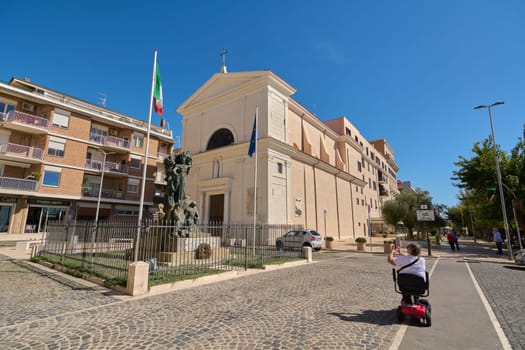 Civitavecchia. Italy - January 27, 2025: The Church of the Japanese Martyrs in Civitavecchia with an ornate facade, prominent statue, flags, and a wheelchair user in the foreground on a sunny day.