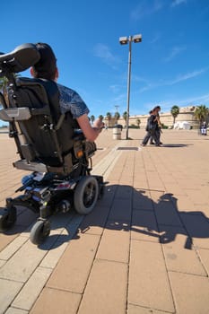 Civitavecchia. Italy - January 27, 2025: January 27, 2025: A wheelchair user enjoys a sunny day on a palm tree-lined promenade. The scene showcases accessibility, leisure, and a clear blue sky.