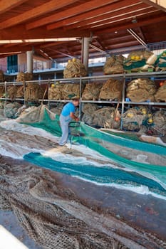 Civitavecchia, Italy - January 31, 2025: In a warehouse full of fishing nets, a fisherman concentrates on repairing them. It highlights the meticulous work and organization of the fishing industry.