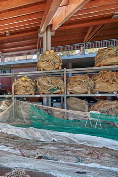 A fishing net repair station in a spacious maritime facility. Large piles of nets, buoys, and ropes are carefully arranged under a wooden-roofed workspace.