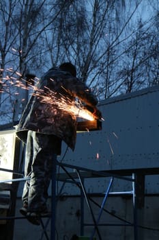 worker with a cutting machine in his hands on a scaffolding cuts metal, a sheaf of bright sparks scatter in the twilight low angle of view