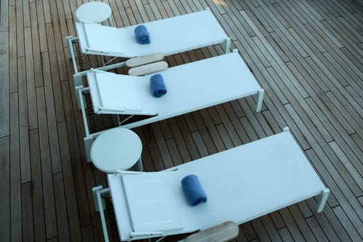 Relaxation. Three white sun loungers with neatly rolled blue towels on a wooden deck. A peaceful outdoor setting ideal for travel, leisure, or wellness concepts.