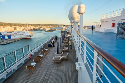 Civitavecchia, Italy - February 6, 2025: Passengers relaxing on the lower deck of a cruise ship with wooden flooring and seating areas, enjoying the serene view of the port city of Civitavecchia.