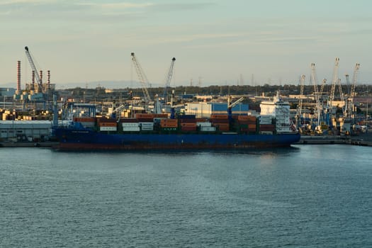 Civitavecchia. Italy - February 06, 2025: A large cargo ship docked at a busy industrial port, loaded with colorful containers and surrounded by cranes and industrial buildings.