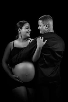 Black and white studio shot of a young African American couple. The woman is pregnant and her belly is visible. They are both looking at each other with a happy expression on their faces.