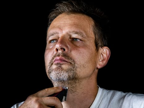 A middle-aged man cuts his gray beard with a shaving trimmer. Mustache and beard clipper. Adult. Male hairdresser. Man's face. Aged people. Hairdresser's tool. Facial care.
