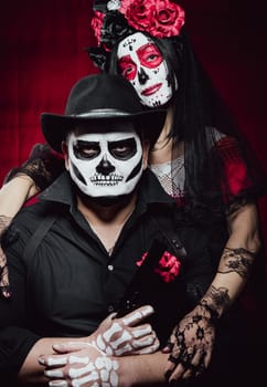 beautiful woman with a sugar skull makeup with a wreath of flowers on her head and a skeleton man in a black hat holding a gun