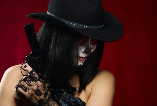 beautiful woman in skeleton make-up holds a revolver, she is wearing a black hat, red background