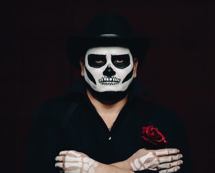 a man is painted with a skeleton in a black hat and shirt, arms crossed on his chest. Black background