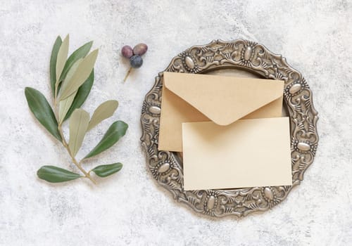 Wedding invitation card and envelope on a plate on a white marble table with olive branches top view. Elegant template with blank paper card flat lay. Mediterranean mockup, place for text