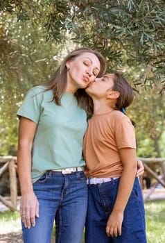 Mother and daughter embracing each other and smiling under olive trees in a park. Cute kid girl and middle aged woman having funoutdoor in summer. Family outdoor activity concept, authentic childhood moment