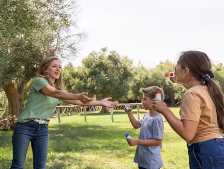 Mother, son and daughter blowing soap bubbles outdoors. Happychildren and middle aged woman having fun in a park in summer. Family and children outdoor activity concept, authentic childhood moment