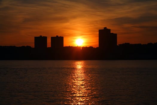 The orange and round sun going behind the skyscrapers and illuminating the Hudson river in New York City