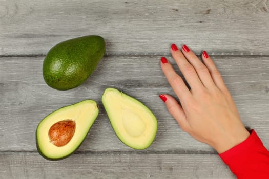 Table top view - avocado cut in half, woman hand with red nails next to it, placed on gray wooden desk.