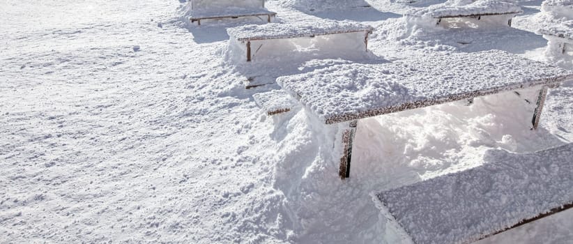 Winter snow loadings banner. Sun shining on desks and benches completely covered under deep snowfall.
