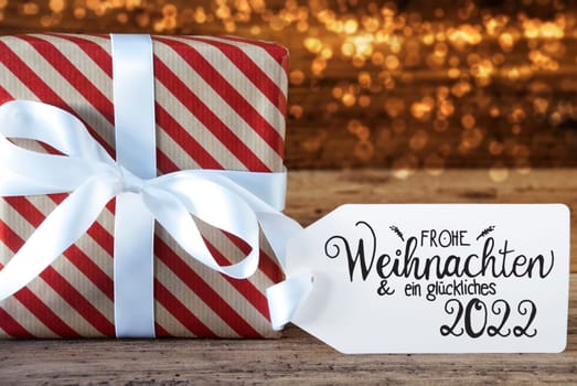 One Christmas Gift With Label With German Calligraphy Frohe Weihnachten Und Ein Glueckliches 2022 Means Merry Christmas And A Happy 2022. Wrapping Paper With A Bow And Lights In Background