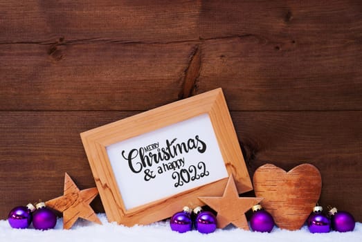 Frame With English Calligraphy Merry Christmas And A Happy 2022. Purple Christmas Decoration Like Ball, Star And Heart. Wooden Background With Snow